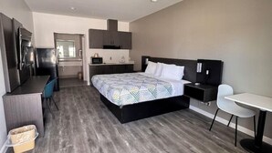 A modern hotel room with a large bed, a desk, a chair, a small table, and a kitchenette area. - Studio6 Humble, TX (Humble)
