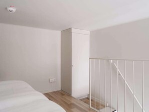 Una habitación con cama, armario, barandilla blanca y detector de humo en el techo.