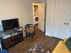 A living room with a television on a wooden stand, a bookshelf, a chair, a door, and a patterned rug. - Center of the action - Family and Pet Friendly (Apex)