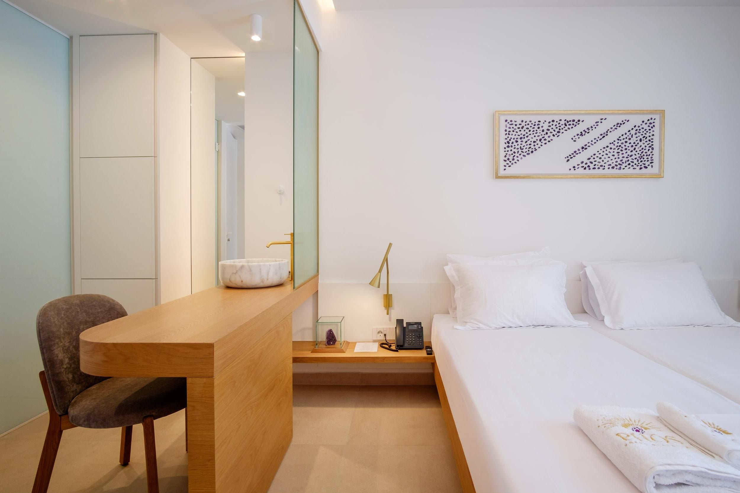 A hotel room with a bed, a desk, a chair, a bedside table with a lamp, and a framed abstract art piece on the wall.