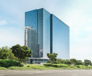 Un edificio moderno de gran altura con fachada de vidrio, rodeado de vegetación y un cielo despejado.