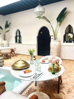 A dining area with a round table set for two, a pool in the background, and a large arched doorway. - Riad El Khaeir Hammam & Spa (Marrakech)