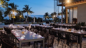 Un espace de restauration en plein air, avec des tables et des chaises, offrant une vue sur un plan d’eau et des palmiers.