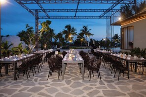 An outdoor dining area with tables set for a meal, surrounded by chairs and palm trees, under a metal structure. - Diamond House Resort Mui Ne (Mui Ne)