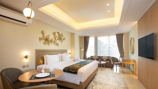 A hotel room with a large bed, a desk, a chair, a TV, and a window with curtains. - Louis Kienne Reserve Cikarang (Cikarang)