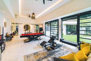 Un salon moderne doté d’une table de billard, d’un espace détente avec un canapé et des fauteuils, et de grandes fenêtres offrant une vue sur l’extérieur.