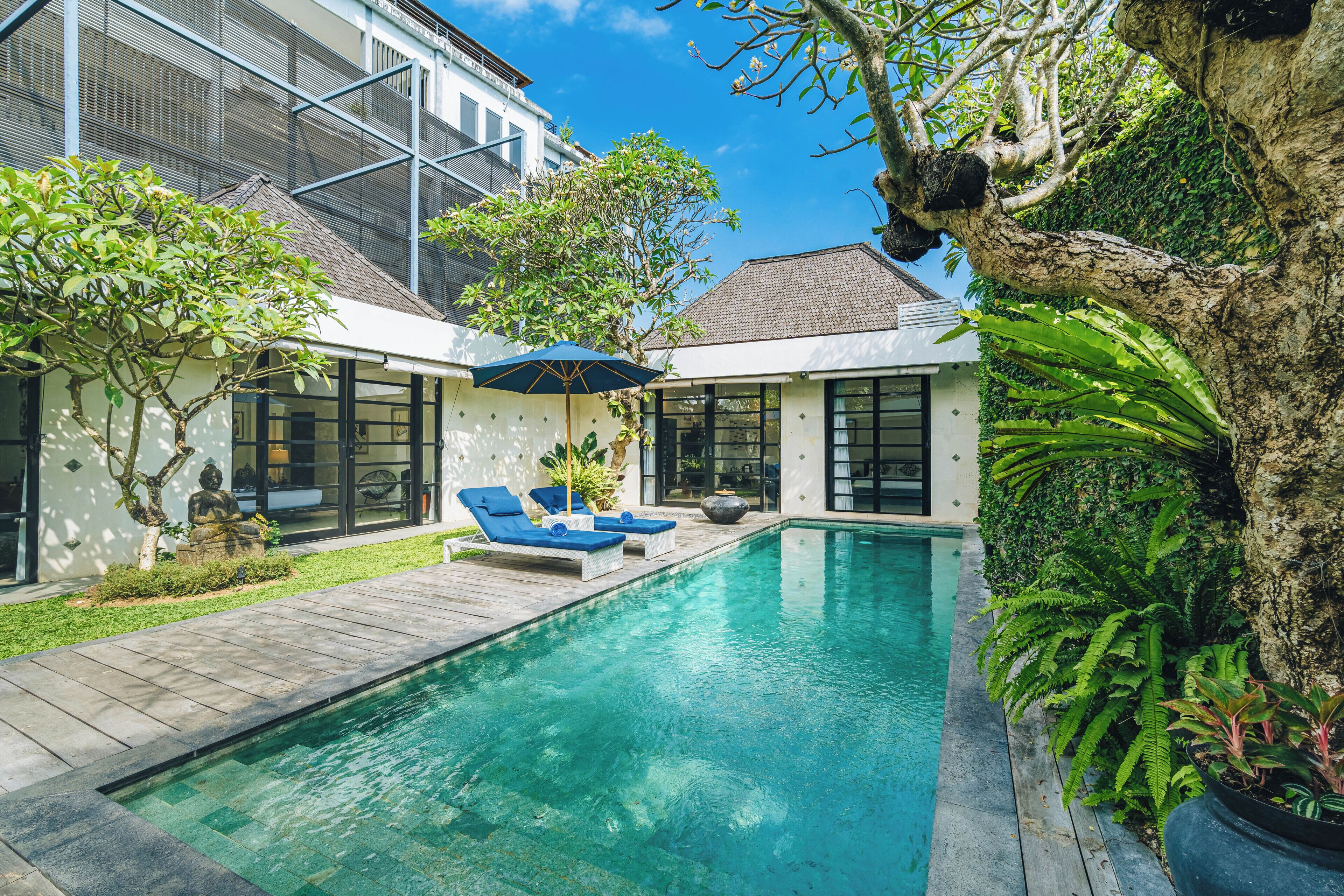 A swimming pool with lounge chairs and a pergola, surrounded by greenery and a building with large windows.