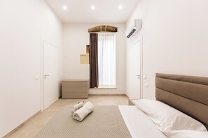 Ein schlicht eingerichtetes Schlafzimmer mit Bett, Nachttisch, kleiner Kommode und einem Fenster mit Vorhängen.