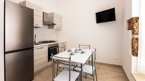 Eine kompakte Küche mit Kühlschrank, Herd, Backofen und Mikrowelle. Ein Esstisch mit Stühlen und ein an der Wand befestigter Flachbildfernseher sind ebenfalls vorhanden.