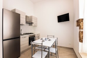 A compact kitchen with a refrigerator, stove, oven, and microwave. There is a dining table with chairs and a flat-screen TV mounted on the wall. - Incanto Holiday House - Appartamento 2 (Avola)