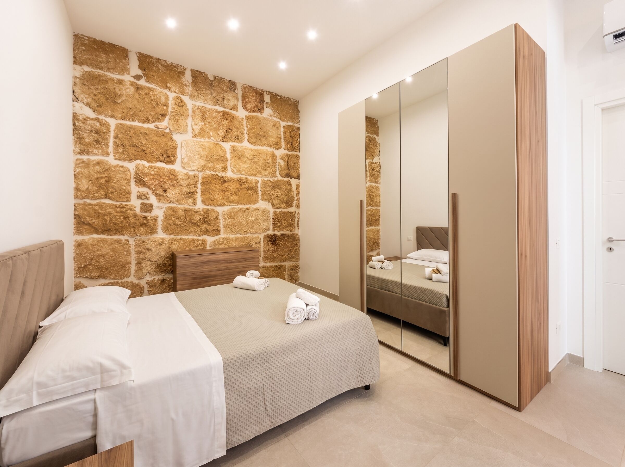 Ein Schlafzimmer mit Steinwand, einem Bett mit weißer Bettwäsche, einem Holzschrank und einem großen Spiegel.