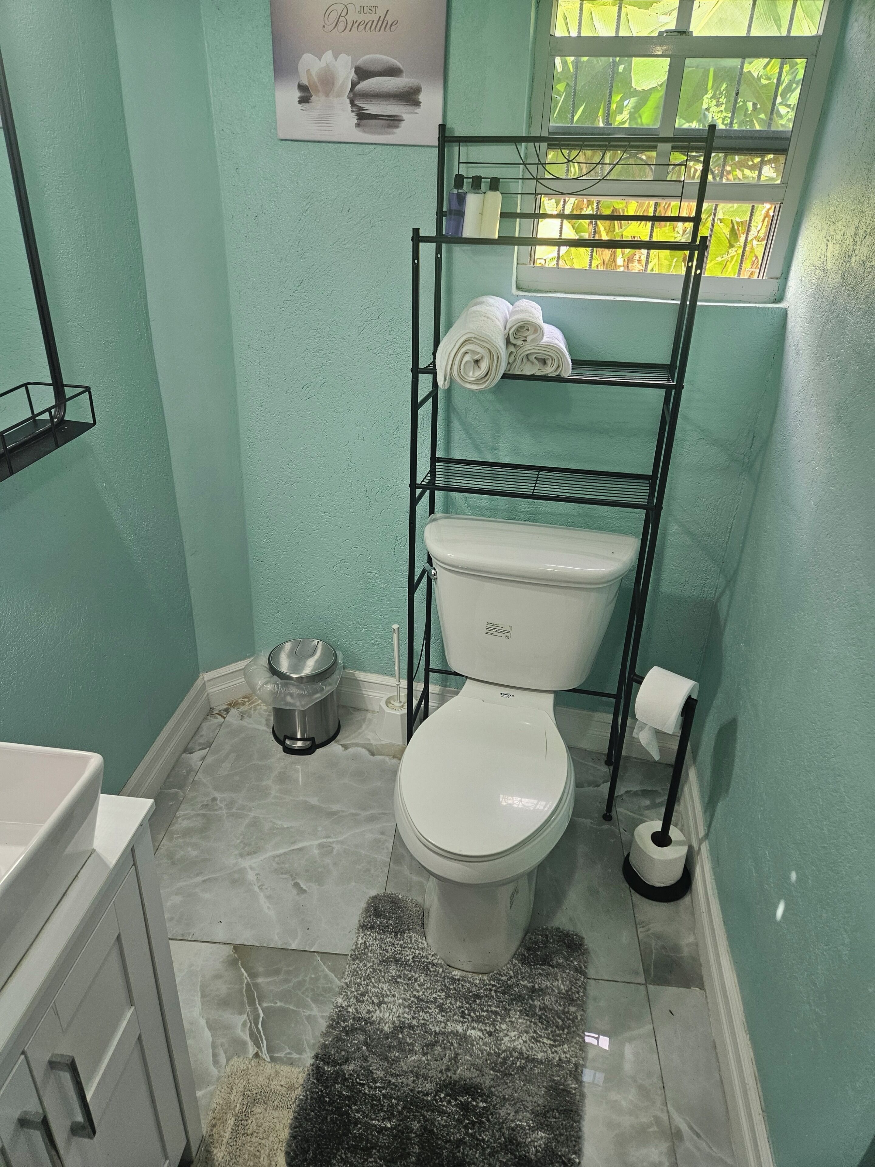 Une salle de bain avec des toilettes, une étagère avec des serviettes, une poubelle et une fenêtre donnant sur un espace verdoyant.