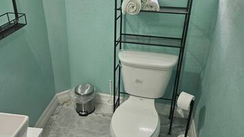Une salle de bain avec des toilettes, une étagère avec des serviettes, une poubelle et une fenêtre donnant sur un espace verdoyant.