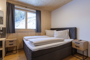 Ein Schlafzimmer mit Bett, Nachttisch, Kommode, einem Fenster mit Vorhängen und Blick auf eine verschneite Landschaft.