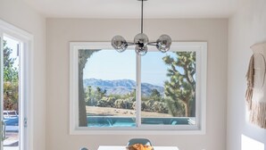 Ein Essbereich mit Tisch, Stühlen, einer Hängeleuchte und Blick auf Pool und Berge durch ein großes Fenster.