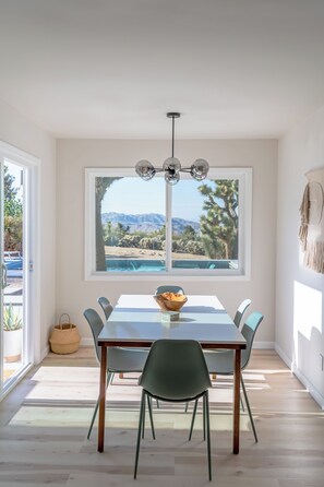 A dining area with a table, chairs, a hanging light fixture, and a view of a pool and mountains through a large window. - Juniper & Sage Retreat - 3 bed, Pool, Spa & Stargazing Near Joshua Tree (Joshua Tree)