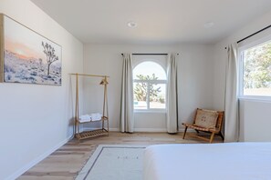 Une chambre à coucher comprenant un lit, une chaise, un étendoir à linge suspendu, une fenêtre avec des rideaux et un tableau accroché au mur.