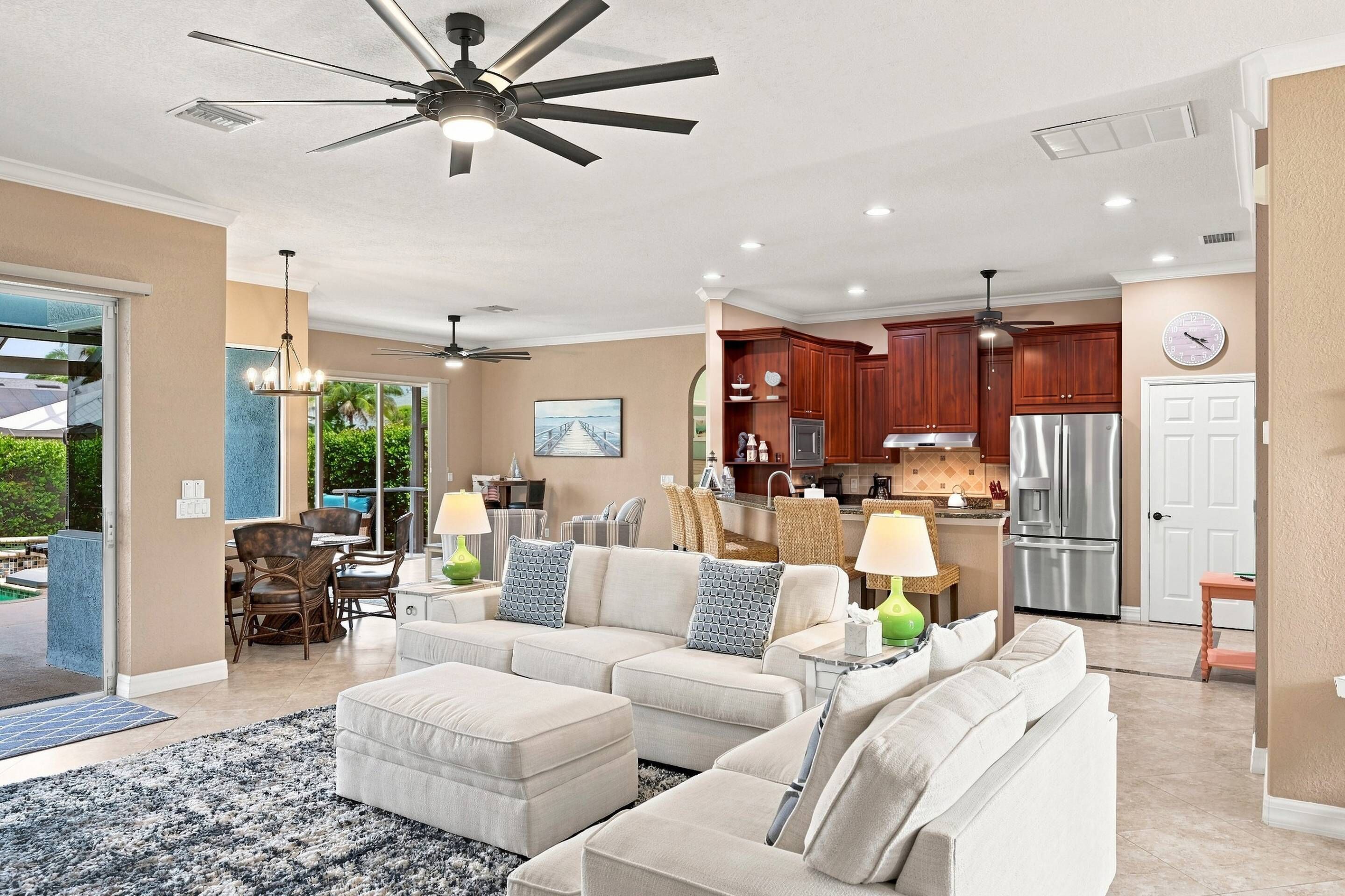 A spacious living room with a large sofa, a dining area with chairs, a kitchen with wooden cabinets, and a ceiling fan.