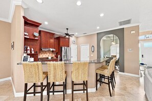 A kitchen with wooden cabinets, a stainless steel refrigerator, and a bar area with woven chairs. - Villa Cecilia | Spacious Cape Coral Escape | Private Heated Pool, Spa, Outdoor Living & Home Office (Cape Coral)