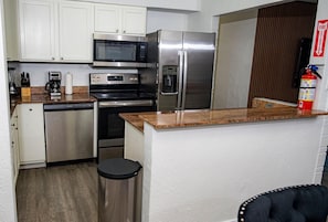 Cocina compacta con armarios blancos, refrigerador de acero inoxidable, microondas, cafetera y basurero.
