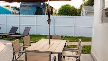 An outdoor patio with a table and chairs, a large umbrella, and a white fence in the background.