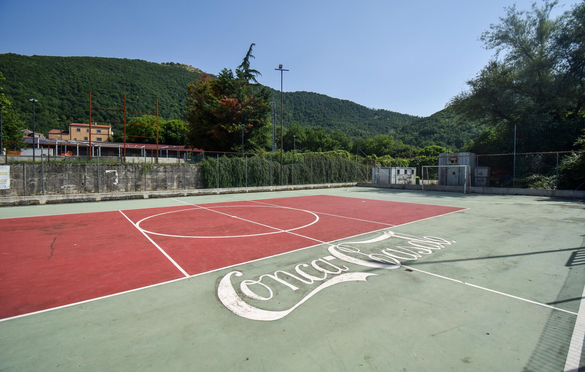 Una cancha de baloncesto al aire libre con el logotipo de Coca-Cola, rodeada de una valla y árboles.