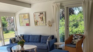 Ein Wohnzimmer mit einem blauen Sofa, einem Korbstuhl, einem runden Tisch und Blick auf Grünflächen.