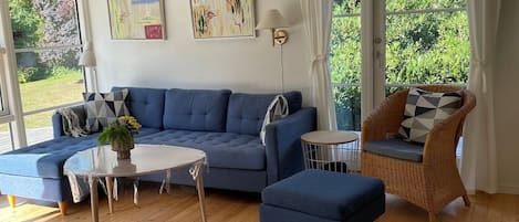 A living room with a blue sofa, a wicker chair, a round table, and a view of greenery outside.
