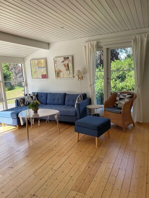 Ett vardagsrum med en blå soffa, en fåtölj i rotting, ett runt bord och utsikt över grönskan utanför.