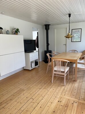 Ett modernt vardagsrum med trägolv, ett matbord med stolar, ett vitt skåp, en svart öppen spis och ett tak med ett rutmönster.