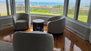 Ein Zimmer mit zwei Sesseln, einem kleinen runden Tisch und großen Fenstern mit Blick auf das Meer und viel Grün.