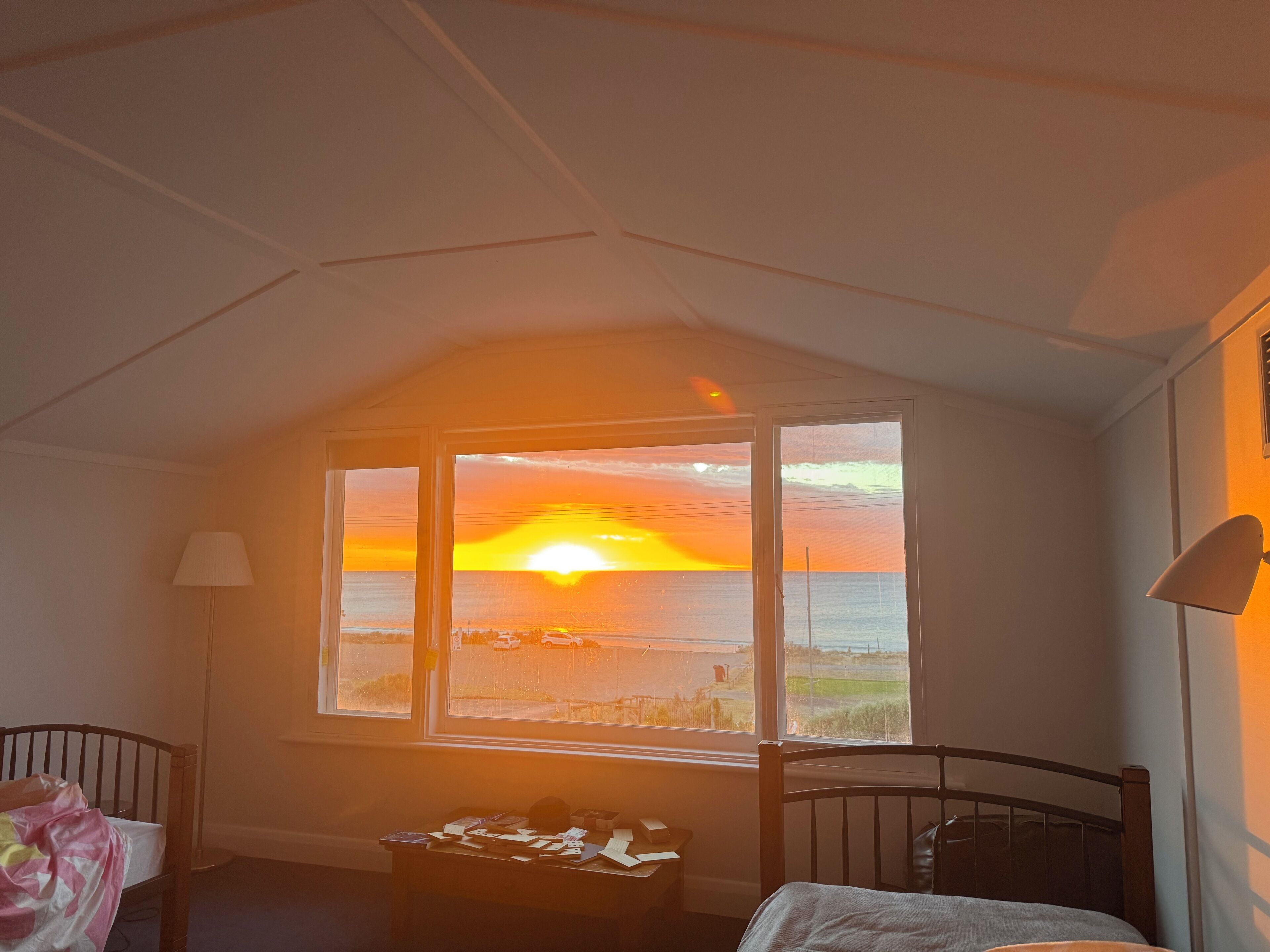 Ein Zimmer mit großem Fenster und Blick auf einen Sonnenuntergang, ein Bett, ein Schreibtisch mit Unterlagen und eine Lampe.