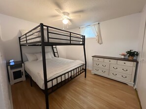 Una cama litera con una mesita de noche, un aparador con cajones, un ventilador de techo y una ventana con cortinas.