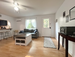 A modern living room with a grey sofa, a coffee table, a TV stand, and a kitchen area with a counter and stools. - Liberty Lounge (Saint Robert)