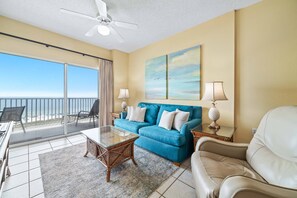 A living room with a blue sofa, a glass coffee table, a white armchair, and a ceiling fan. - Tidewater 608 | Indoor/Outdoor Pools + Hot Tub, Fitness & Sauna (Orange Beach)
