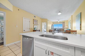 A kitchen with a white countertop, stainless steel sink, and a dishwasher. There are framed pictures on the wall, a ceiling fan, and a view of the living area through an open door. - Tidewater 608 | Indoor/Outdoor Pools + Hot Tub, Fitness & Sauna (Orange Beach)