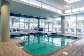 An indoor swimming pool with clear glass walls, lounge chairs, and a tiled floor. - Tidewater 608 | Pools + Hot Tub + Fitness (Orange Beach)