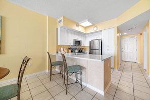 A kitchen with white cabinets, a stainless steel refrigerator, and a microwave. There are two bar stools and a dining table with chairs. - Tidewater 608 | Pools + Hot Tub + Fitness (Orange Beach)