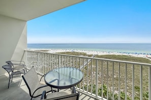 Un balcon avec une table et des chaises en verre, donnant sur une plage parsemée de parasols et fréquentée par des gens.