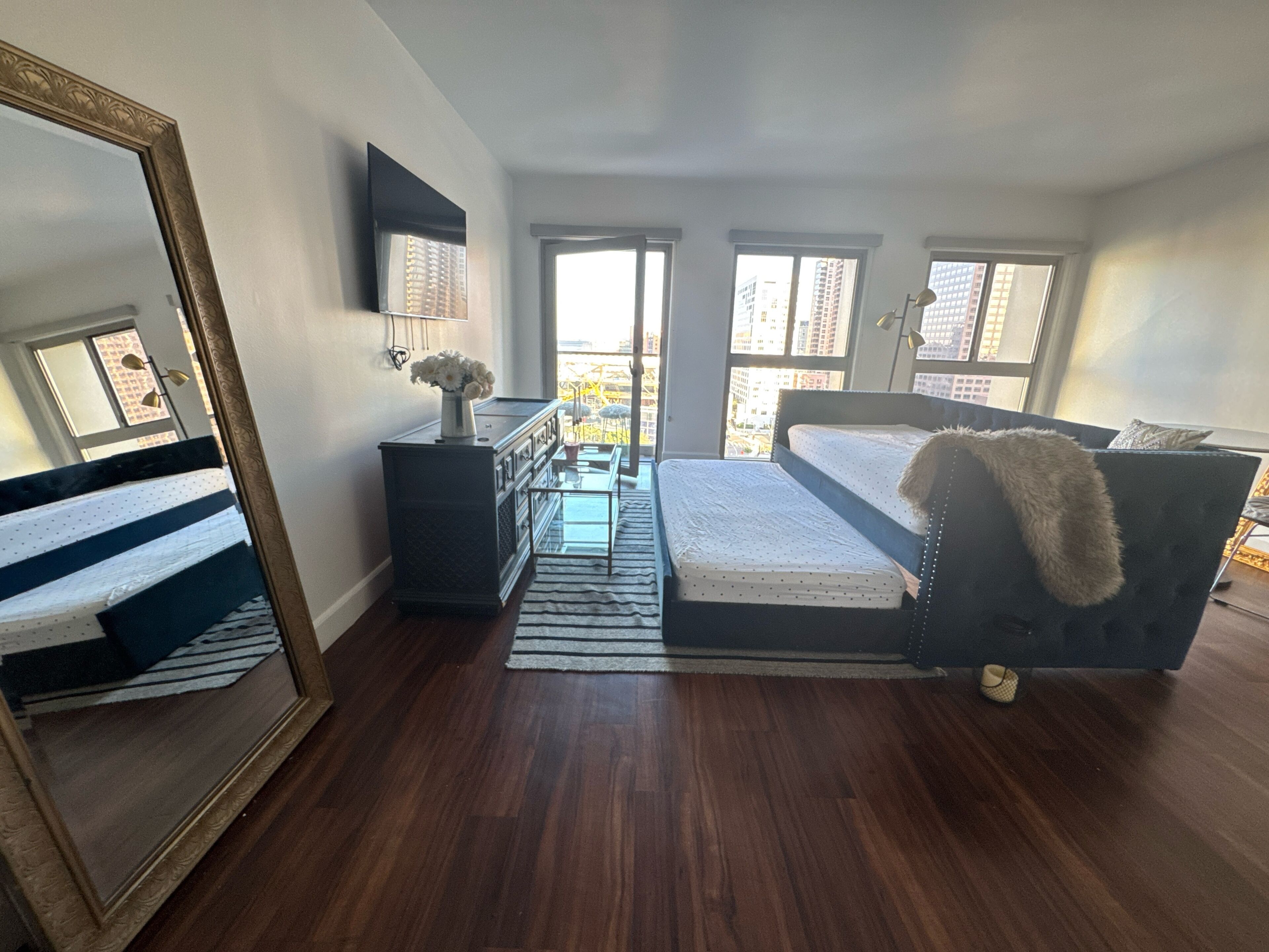 Quarto com cama, cômoda, espelho e janelas com vista para a cidade.
