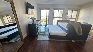 Quarto com cama, cômoda, espelho e janelas com vista para a cidade.