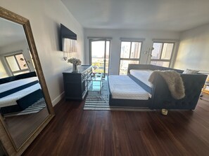 A bedroom with a bed, dresser, mirror, and windows with city views.