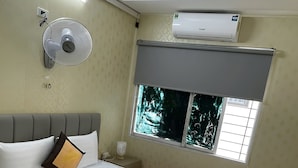 Ett hotellrum med en säng, en fläkt som är monterad på väggen, en luftkonditionering och ett fönster med utsikt över träd.
