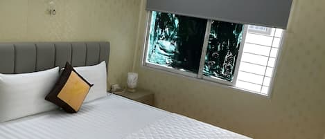 A hotel room with a bed, a wall-mounted fan, an air conditioner, and a window with a view of trees.