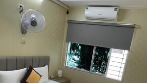 Ett hotellrum med en säng, en fläkt som är monterad på väggen, en luftkonditionering och ett fönster med utsikt över träd.