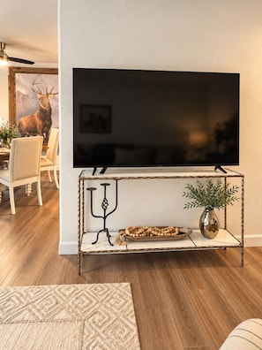 Una sala de estar con un televisor de pantalla plana montado en la pared, una consola de madera con objetos decorativos, y una zona de comedor con una silla y una mesa.