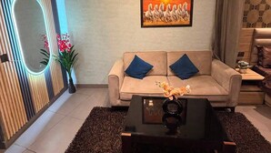 A modern living room with a sofa, a coffee table, a chandelier, a mirror, and a framed picture on the wall. - The Spira Skyline Stay (Noida)