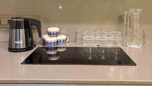 A kitchen with a stainless steel coffee maker, a stack of patterned cups, and a clear glass pitcher on a black induction cooktop. - The Spira Skyline Stay (Noida)