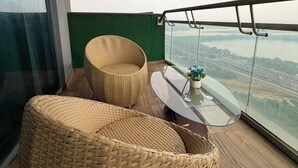 A balcony with wicker furniture, a glass table, and a view of a bridge and water. - The Spira Skyline Stay (Noida)