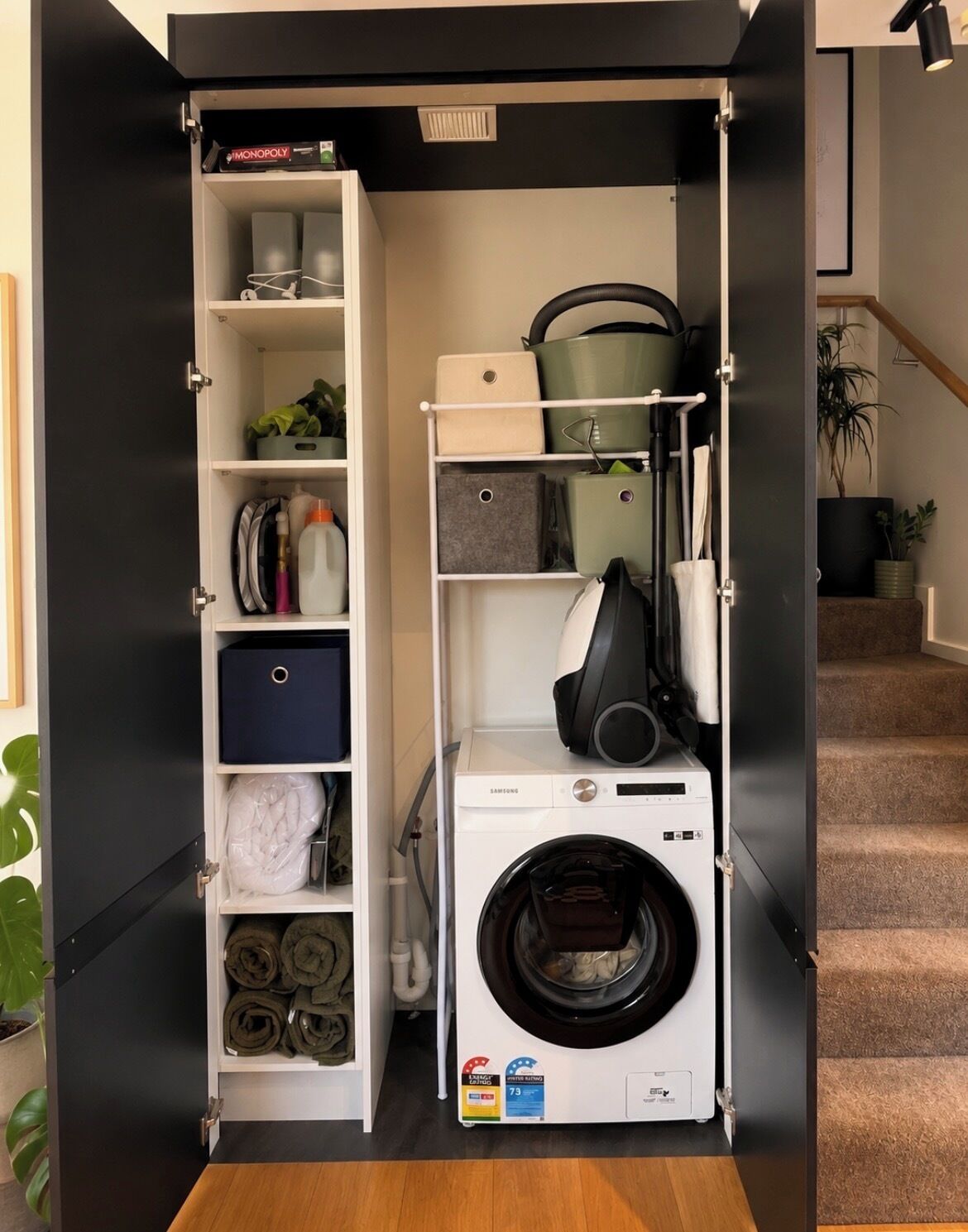A compact laundry room with a front-loading washing machine, shelves with folded clothes, and a shelf with a board game.